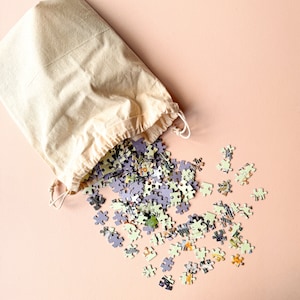 May include: A beige canvas bag with a drawstring closure, spilling out a pile of colorful jigsaw puzzle pieces on a light pink background.