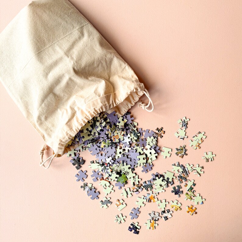 May include: A beige canvas bag with a drawstring closure, spilling out a pile of colorful jigsaw puzzle pieces on a light pink background.