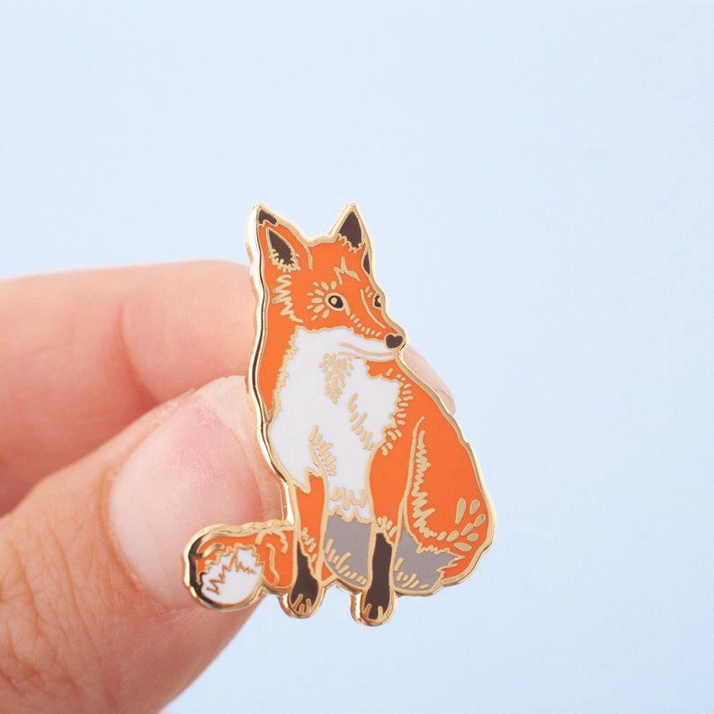 May include: A gold-toned enamel pin featuring a red fox with white fur on its chest and a bushy tail. The fox is sitting with its head turned to the side.