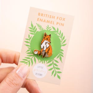 May include: A close-up of a British Fox enamel pin on a card with green foliage. The pin is orange and white with a gold outline. The card reads "British Fox Enamel Pin" and "Little Parsley Designs".