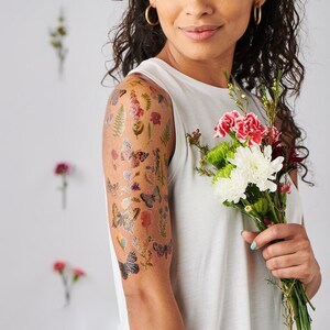 Puede incluir: Un brazo con un tatuaje temporal de mariposas y flores. El tatuaje est&aacute; en la parte superior del brazo y es visible desde el hombro hasta el codo. La persona sostiene un ramo de flores en su mano derecha.