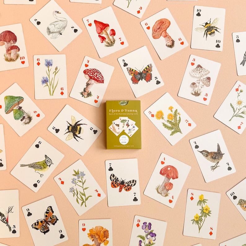 May include: A deck of playing cards featuring illustrations of flora and fauna. The cards are printed on white cardstock with colorful illustrations of mushrooms, butterflies, bees, birds, and flowers. The box for the cards is green and features the text "Flora & Fauna, Deck of Illustrated Playing Cards".