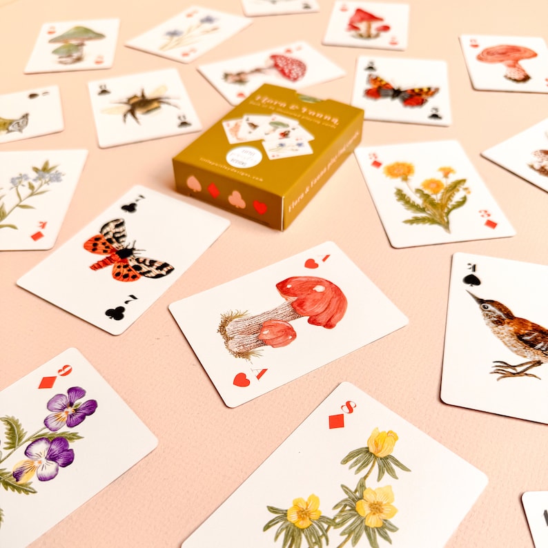 May include: A deck of playing cards with illustrations of flora and fauna. The cards feature images of mushrooms, butterflies, birds, and flowers. The deck is called "Flora & Fauna" and is made by "The Curious Fox Co."