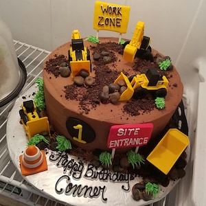 May include: A chocolate cake decorated with construction vehicles, rocks, and green grass. The cake is for a first birthday and says "Happy Birthday Conner" and "Site Entrance" on the side.
