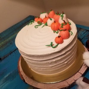 May include: A white cake with buttercream frosting decorated with fondant pumpkins and green vines. The cake is on a wooden cake stand with a gold trim.