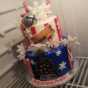 May include: A two-tiered birthday cake with a Polar Express theme. The top tier is white with red stripes and snowflake decorations. The bottom tier is blue with a train design. Candy canes and a bell complete the festive look. The cake reads "Happy Birthday Michelle!"