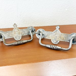 May include: Two antique silver-toned drawer pulls. Each pull features a decorative rectangular backplate with an embossed design and a curved handle. The pulls are resting on a wooden surface.