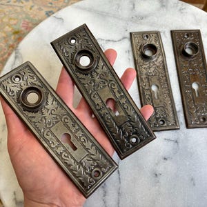 May include: Four rectangular metal door plates with a dark, aged finish. Each plate features an intricate embossed floral and scrollwork pattern, a central circular opening for a door knob, and a lower keyhole cutout with mounting holes.