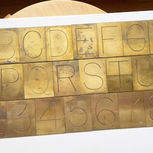 May include: A set of brass letter and number stamps. The stamps are arranged in three rows. The top row contains the letters A through I. The middle row contains the letters O through V. The bottom row contains the numbers 2 through 9.