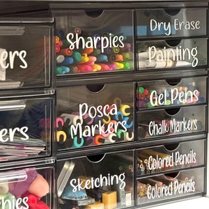 May include: A clear plastic multi-drawer storage unit organized with white labels. Drawers contain various art supplies including Sharpies, Posca markers, gel pens, chalk markers, colored pencils, and sketching tools, with visible text labels on each drawer front.