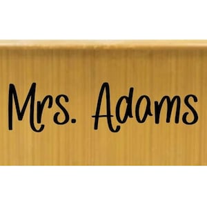 May include: A rectangular desk nameplate with a light brown wood grain finish. The text "Mrs. Adams" is printed in a bold, black, sans-serif font across the center of the front surface. The nameplate has a smooth, flat, horizontal orientation.