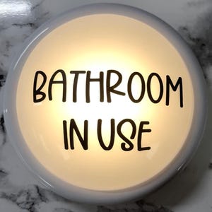 May include: A round, white bathroom sign with the words "BATHROOM IN USE" in black lettering. The sign has a soft, glowing light emanating from within, creating a warm, inviting ambiance. The background is a marble-like pattern.