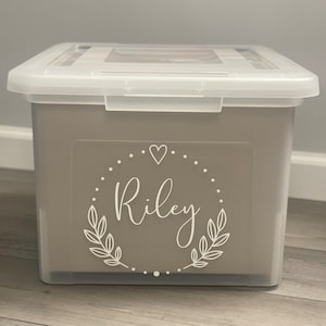 May include: A clear plastic storage bin with a gray lid. The bin has a white label with a heart and the name "Riley" in a decorative font.