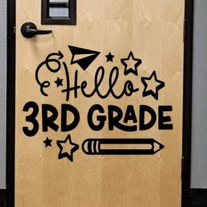 May include: A wooden door with a black door handle and a black framed window. The door features a black decal that says "Hello 3rd Grade" with a paper airplane, stars, and a pencil.