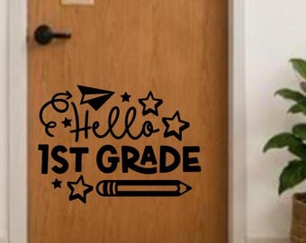 May include: A black vinyl wall decal featuring the text Hello 1ST GRADE in a stylized font. The design includes a paper airplane, several five-pointed stars, and a pencil graphic. The decal is applied to a smooth, light brown wooden door surface.