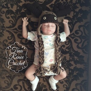 K&ouml;nnte beinhalten: Ein Baby tr&auml;gt eine braune geh&auml;kelte Elch-M&uuml;tze mit langen Z&ouml;pfen. Die M&uuml;tze hat braune Filz-Geweihe und schwarze Knopfaugen. Das Baby tr&auml;gt einen wei&szlig;en und braunen gemusterten Strampler.