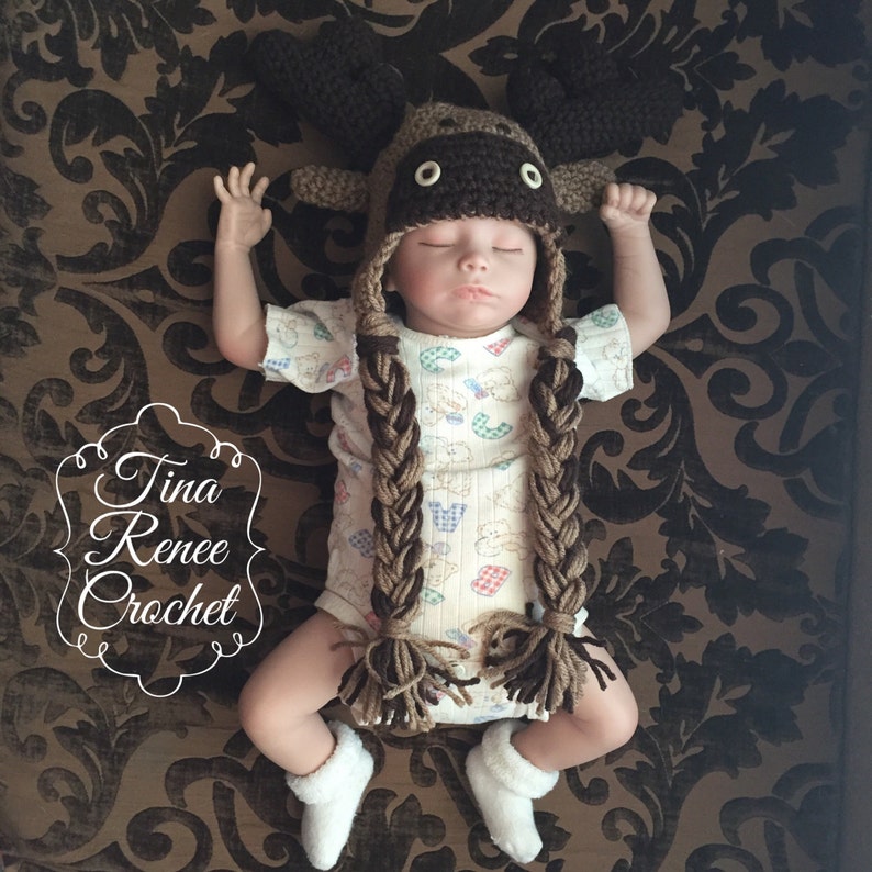 K&ouml;nnte beinhalten: Ein Baby tr&auml;gt eine braune geh&auml;kelte Elch-M&uuml;tze mit langen Z&ouml;pfen. Die M&uuml;tze hat braune Filz-Geweihe und schwarze Knopfaugen. Das Baby tr&auml;gt einen wei&szlig;en und braunen gemusterten Strampler.