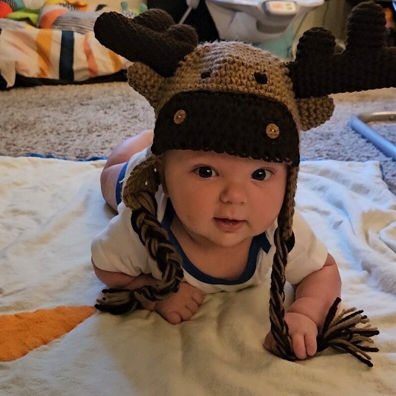 K&ouml;nnte beinhalten: Ein Baby tr&auml;gt eine braune und beige geh&auml;kelte Elch-M&uuml;tze mit langen braunen Z&ouml;pfen. Die M&uuml;tze hat gro&szlig;e Elchenohren und braune Knopfaugen.