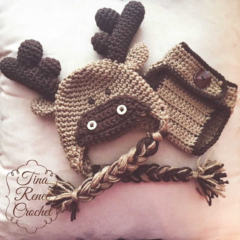 K&ouml;nnte beinhalten: Ein geh&auml;keltes Elch-M&uuml;tzen- und Windelset f&uuml;r ein Baby. Die M&uuml;tze hat ein braunes Gesicht mit wei&szlig;en Augen und einen geflochtenen Schwanz. Die Windel hat eine braune Tasche mit einem Knopf.