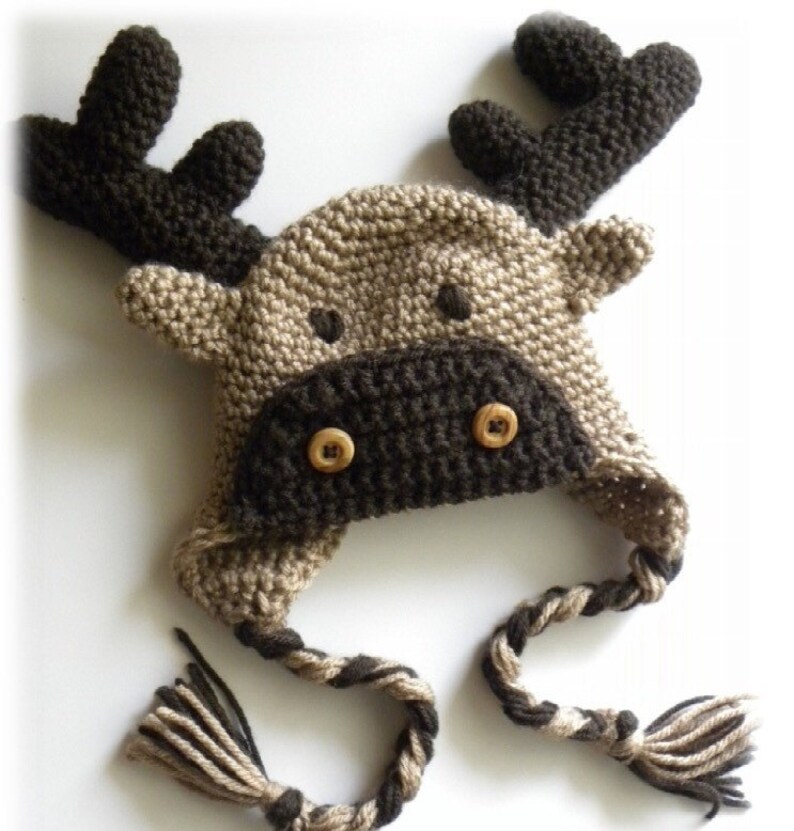 K&ouml;nnte beinhalten: Eine braune und beige gestrickte M&uuml;tzen in Form eines Elches mit einer langen, geflochtenen Schnur. Die M&uuml;tze hat zwei braune Kn&ouml;pfe f&uuml;r die Augen.