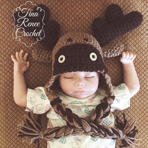 K&ouml;nnte beinhalten: Ein Baby tr&auml;gt eine braune und beige gestrickte M&uuml;tzen mit langen geflochtenen Garnenden. Die M&uuml;tze hat gro&szlig;e braune Ohren und braune Knopfaugen.