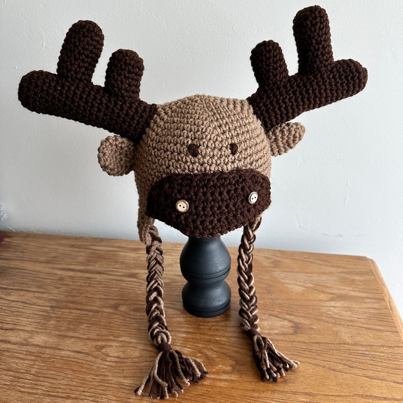 K&ouml;nnte beinhalten: Eine braune und beige gestrickte M&uuml;tzen mit Knopfaugen und langen geflochtenen Quasten.