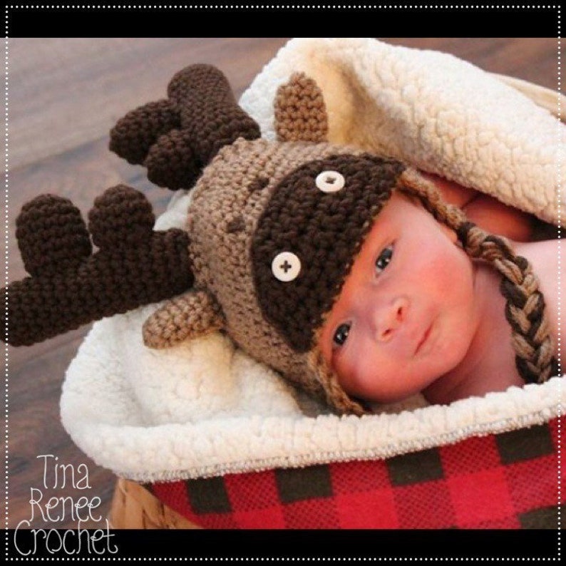 K&ouml;nnte beinhalten: Ein Baby tr&auml;gt eine braune und beige gestrickte Elch-M&uuml;tze mit gro&szlig;en braunen Geweih. Das Baby liegt in einem wei&szlig;en und roten karierten Korb.