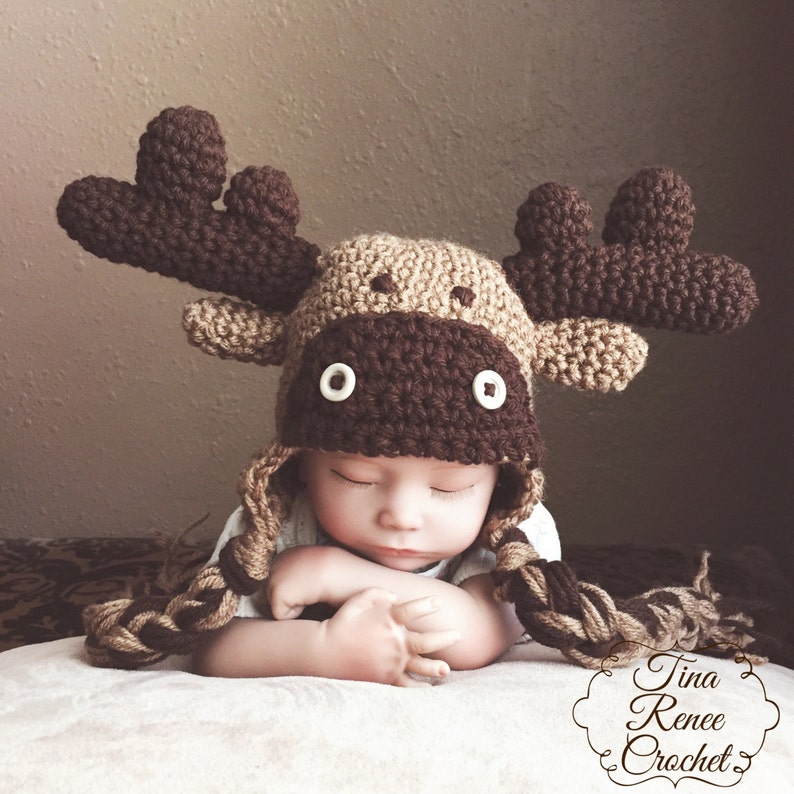 K&ouml;nnte beinhalten: Ein Baby tr&auml;gt eine braune und beige gestrickte M&uuml;tzen mit wei&szlig;en Knopfaugen. Die M&uuml;tze hat lange braune und beige gestrickte Z&ouml;pfe.