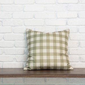 May include: A square throw pillow featuring a classic gingham pattern in muted sage green and off-white. The fabric has a soft, matte texture with a consistent grid design covering the entire front surface of the cushion, creating a balanced look.