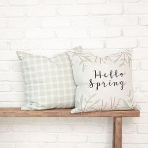 Puede incluir: Dos cojines cuadrados. Uno presenta un patrón de cuadros vichy en verde claro y blanco. El otro es de color blanco roto con un diseño estampado de ramas finas marrones y hojas verde claro, con el texto Hello Spring en color negro.