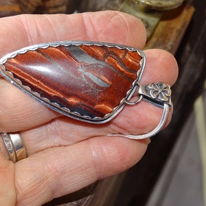 May include: A silver pendant with a red and brown tiger's eye stone set in a scalloped bezel. The pendant is attached to a silver chain with a flower-shaped clasp.