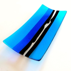 Puede incluir: Un plato rectangular de vidrio con forma curva. El plato presenta un degradado de tonos azules, con una franja diagonal negra y blanca. El plato probablemente se utiliza con fines decorativos o como una pequeña bandeja de servicio.