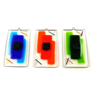 May include: Three rectangular glass ornaments with a white background and colourful geometric shapes. Each ornament features a small black square in the centre and a silver hanging loop.