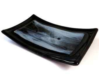 May include: A black glass soap dish with a white swirl design. The dish has a curved shape and a small drainage hole.
