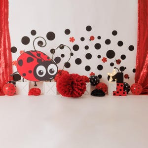 May include: A red and black ladybug cutout with white polka dots, a red pom-pom, and a white wooden block with the number "1" on it. The background is white with black polka dots and red curtains.