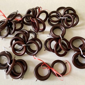 May include: A collection of dark brown wooden curtain rings, some bundled with white or red ribbons. Each ring has a small metal eyelet. The rings are arranged on a light-colored surface.