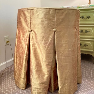 May include: A gold-colored, silk-like table skirt with pleats and button details. The skirt covers the entire table, creating a full, elegant look. The fabric has a subtle sheen, adding to its luxurious appearance.