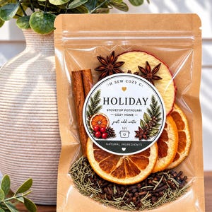 May include: A brown kraft paper pouch containing stovetop potpourri with dried orange slices, cinnamon sticks, star anise, cloves, and rosemary. The circular label reads Oh Sew Cozy Co Holiday Stovetop Potpourri Cozy Home Just Add Water.