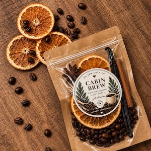 May include: A clear plastic pouch containing stovetop potpourri labeled Cabin Brew, featuring dried orange slices, coffee beans, a cinnamon stick, and a vanilla bean. The label includes illustrations of pine trees and a coffee cup.