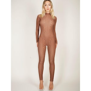 Może przedstawiać: Brązowy kombinezon z wzorem słojów drewna. Kombinezon ma długie rękawy i wysoki dekolt. Modelka ma na sobie przezroczyste obcasy. Strój jest dopasowany i zakrywa całe ciało.