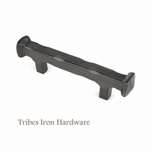 May include: A dark brown, rustic-style cabinet pull with a textured, hammered appearance. The handle is rectangular with two square posts and square ends. The text "Tribes Iron Hardware" is below the pull.