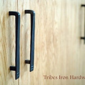 Könnte beinhalten: Drei schwarze Metall-Schrankgriffe mit rechteckiger Form und leicht abgerundeter Oberseite. Die Griffe sind auf einer hellbraunen Holzoberfläche montiert. Der Text "Tribes Iron Hardware" ist am unteren Rand des Bildes sichtbar.
