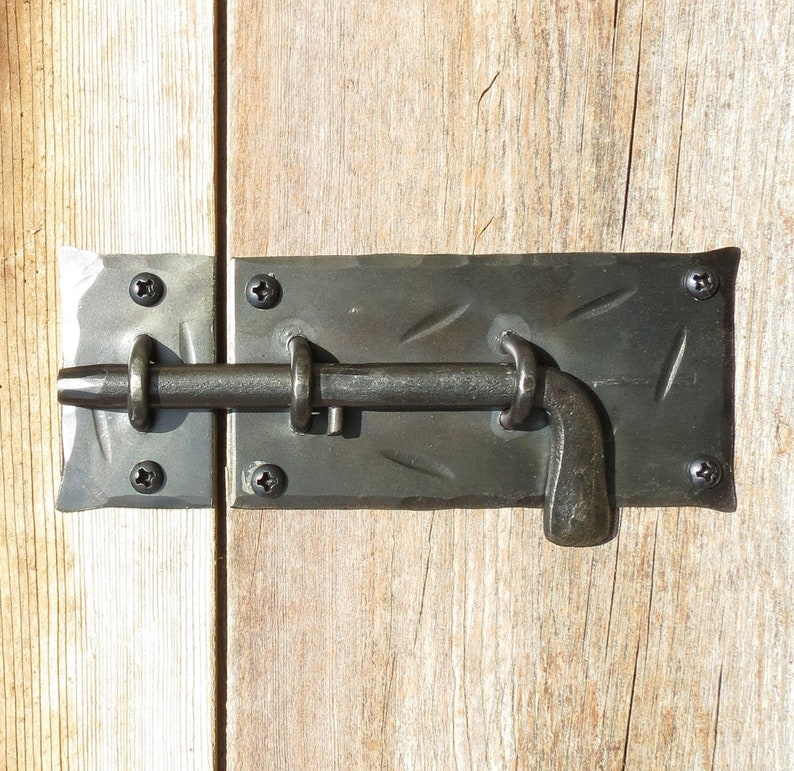 Puede incluir: Un pestillo de puerta de metal negro con un dise&ntilde;o decorativo. El pestillo tiene una placa rectangular con una manija curva y un perno que se desliza a trav&eacute;s de la placa.