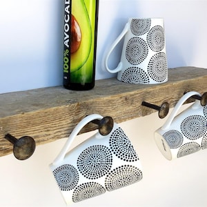 May include: Rustic wooden shelf with four metal hooks, each holding a white mug with a black and white circular pattern. A bottle of 100% avocado oil is on the shelf.