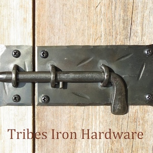 Puede incluir: Un pestillo de puerta de metal negro con una superficie texturizada y una manija curva. El pestillo est&aacute; fijado a una puerta de madera con tornillos. El texto "Tribes Iron Hardware" es visible debajo del pestillo.