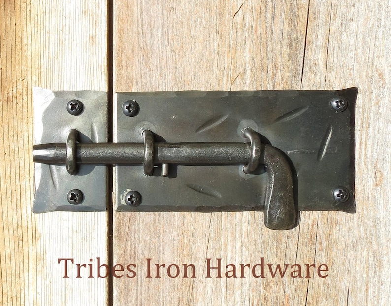 Puede incluir: Un pestillo de puerta de metal negro con una superficie texturizada y una manija curva. El pestillo est&aacute; fijado a una puerta de madera con tornillos. El texto "Tribes Iron Hardware" es visible debajo del pestillo.