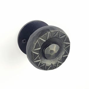 Può includere: Un pomello per porta nero, strutturato, con un design circolare. Il pomello presenta un elemento centrale sfaccettato circondato da un motivo di forme triangolari. Il pomello è montato su una piastra posteriore rotonda nera con una singola vite.