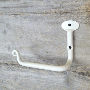 May include: A white, hand-forged metal toilet paper holder. The holder has a curved arm with a decorative swirl at the end and an oval mounting plate. The holder is mounted with two black screws and is designed for wall mounting.