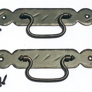 May include: Two black metal drawer pulls with a decorative hammered finish and a diamond pattern. Each pull has a rounded rectangular base with a pointed end on each side. The pulls have a simple loop handle.