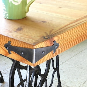 May include: A rustic coffee table with a light-colored wooden top and black metal corner brackets. The table is supported by a vintage black cast iron base, likely from a sewing machine. A green mug with a white floral design sits on the table.